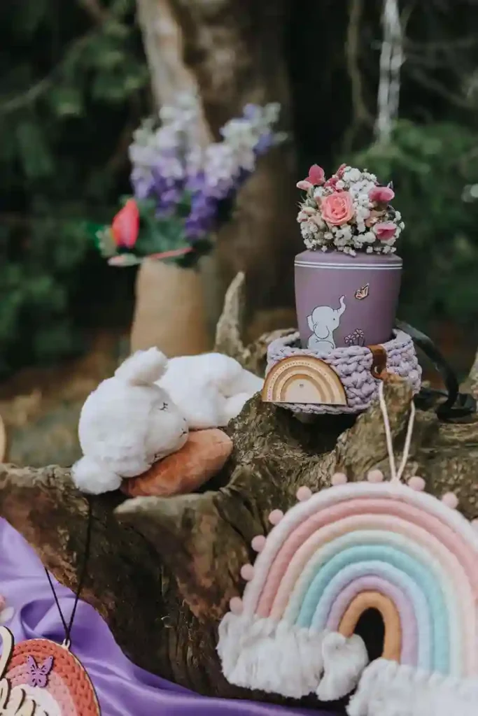 Sternenkind-Urne in zartem Violett mit Blumenarrangement, umgeben von Stofftier und Regenbogen-Dekoration als liebevolle Erinnerung
