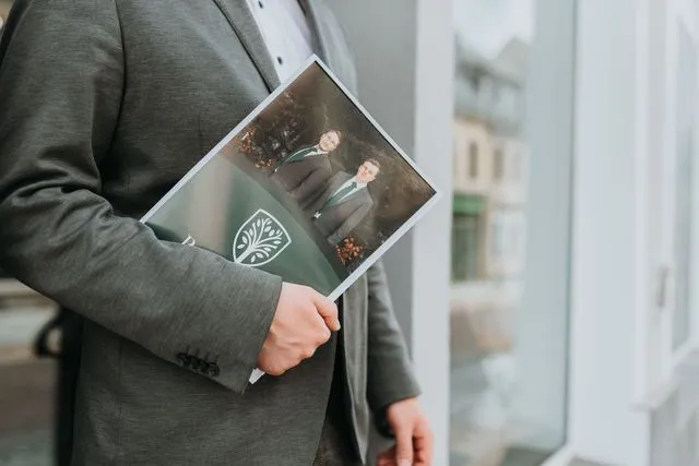 Bestatter hält eine Informationsmappe mit Portrait der Inhaber vor dem Eingang des Bestattungshauses Paul & Baar